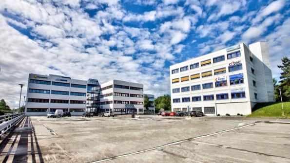 Moderne kontorbygg med parkeringsplass og blå himmel i bakgrunnen.