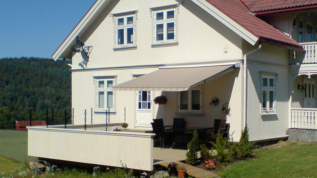 Moderne hus med terrasse, markise og hage, omgitt av grønt landskap.