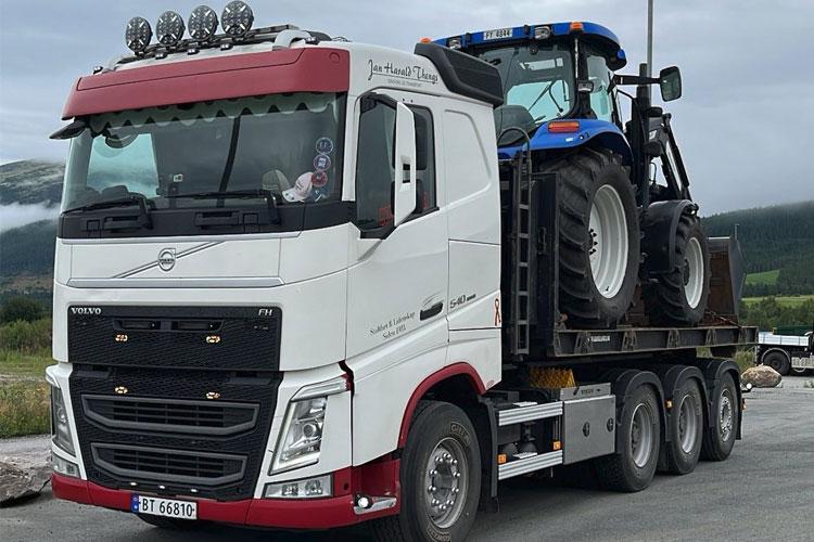 En stor lastebil med en blå traktor på tilhenger, parkert i et naturskjønt område.
