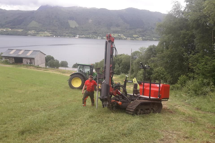 En mann står ved en gravemaskin på en grønn eng med en traktor i bakgrunnen og en innsjø i horisonten.