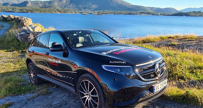 En svart Mercedes SUV parkert ved en innsjø med fjell i bakgrunnen og klart blått vann.
