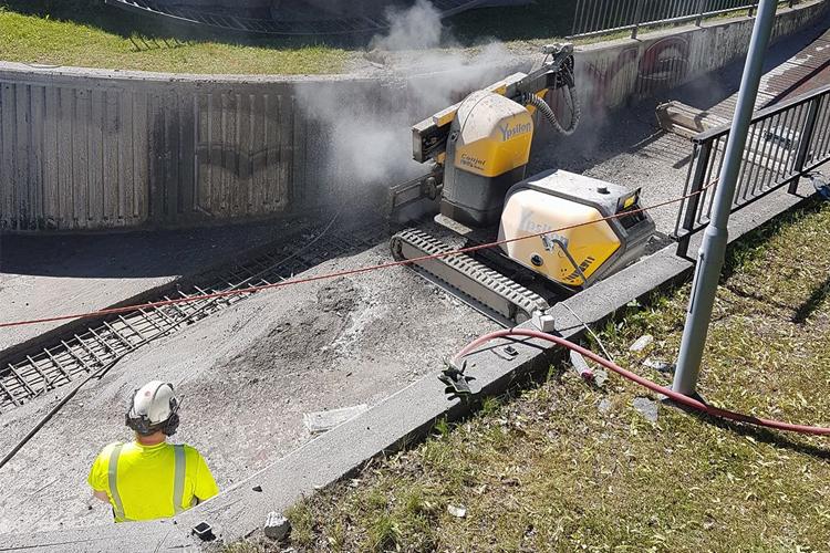 En arbeider i sikkerhetsutstyr ser på en betongskjærer som jobber på en byggeplass. Røyk og støv er synlig.