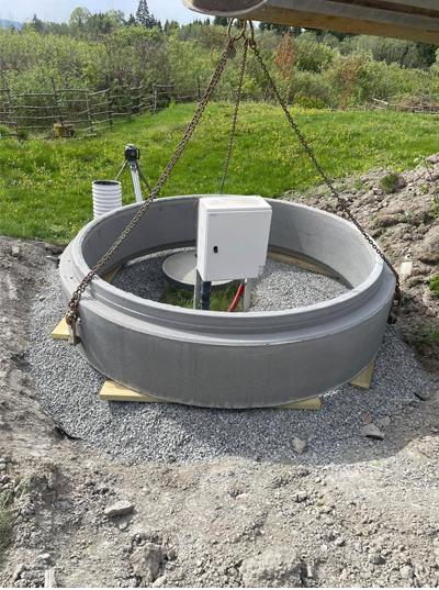 En stor betongring med en hvit boks i midten, plassert på grus under en heis. Grønne områder i bakgrunnen.