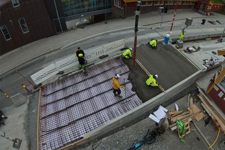 Arbeidere legger betong på en byggeplass, med armeringsjern og verktøy synlig rundt dem.