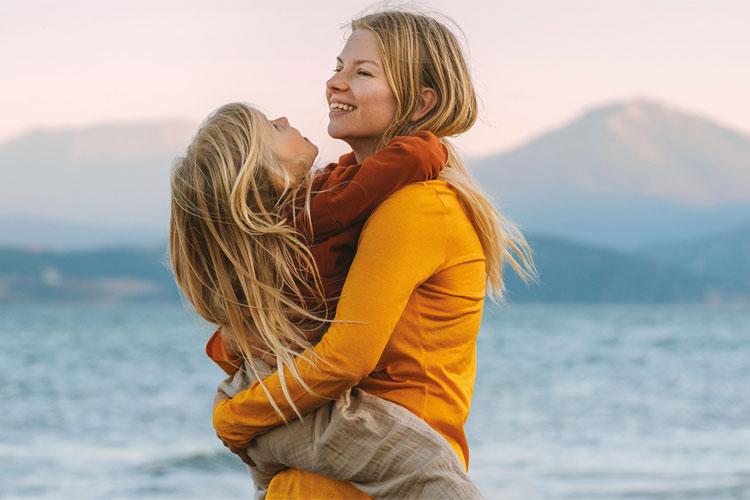 Mor og datter klemmer hverandre på stranden med fjell i bakgrunnen, begge smilende og glade.