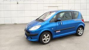 Blå liten bil parkert foran en hvit vegg, med aluminiumsfelger og en sporty design.