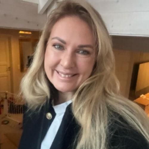 En kvinne med langt, lyst hår smiler mot kameraet i et lyst rom med moderne interiør.
