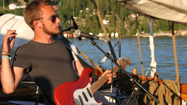 Mann med solbriller spiller elektrisk gitar på en seilbåt ved vannet, med landskap i bakgrunnen.
