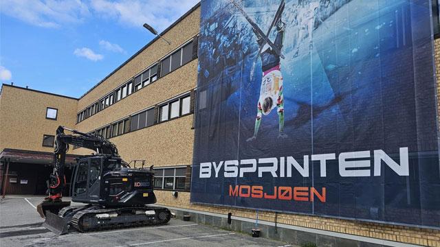 Gravemaskin parkert foran en stor plakat med en svømmer i aksjon, ved Bysprinten i Mo i Rana.
