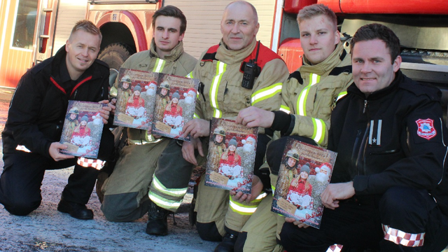 Firemenn holder julekalendere foran brannbilen, iført brannuniformer og smilende mot kameraet.