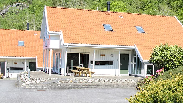 Moderne hytte med oransje tak, terrasse og grønne planter i hagen, omgitt av frodig natur.