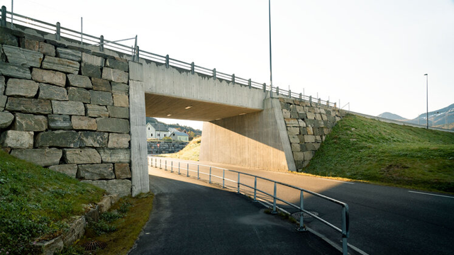 Betongbro med steinvegg og vei som går under, omkranset av grønt landskap og fjell i bakgrunnen.