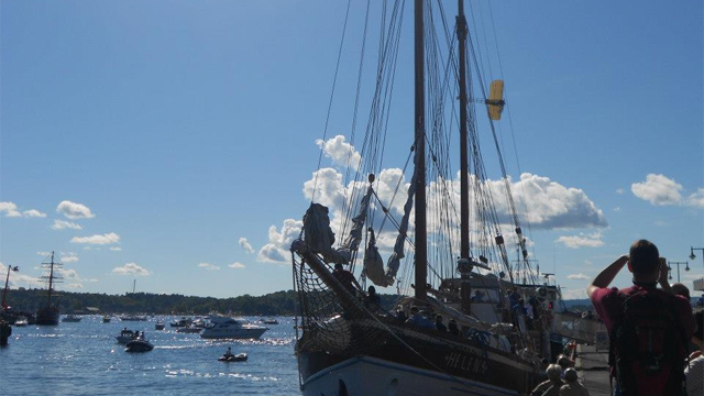 Seilbåt ved havnen med folk som tar bilder, blå himmel og skyer i bakgrunnen.