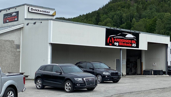 Bilverksted med to svarte biler parkert foran, beliggende i naturskjønt område. Skilt med "Andersen Bil og Dekkservice AS".