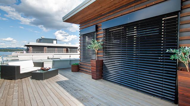 Moderne terrasse med tredekk, svarte persienner og grønne planter, med utsikt over vannet.
