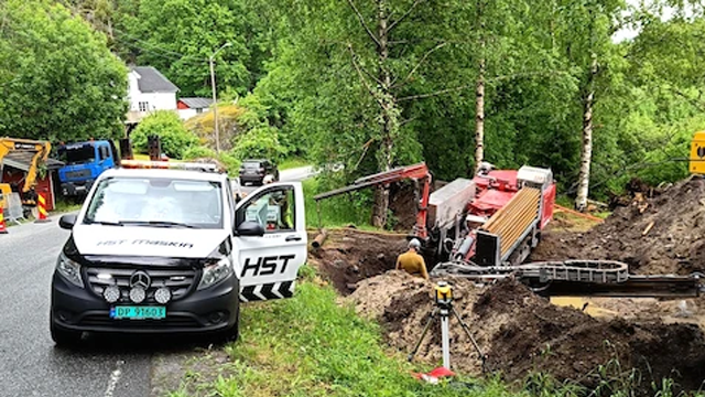 En arbeidsbil fra HST står parkert ved en vei med gravemaskin og arbeidere i en grøft.