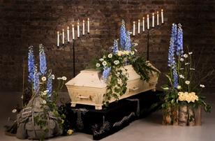 En kiste dekorert med blomster og lys, omgitt av blå delphinium og hvite blomster i et rom med murvegg.