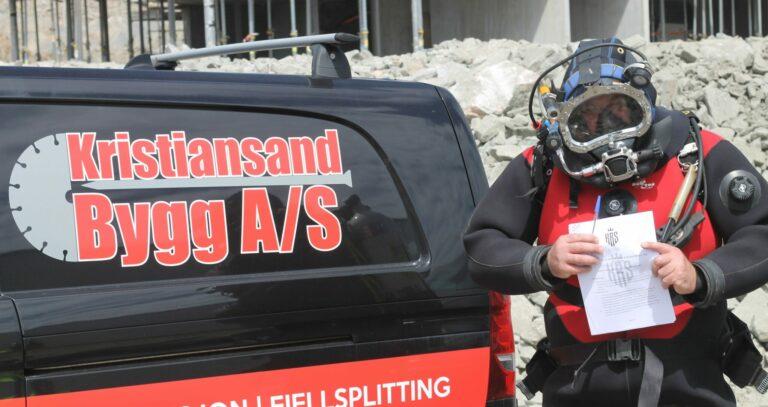 Dykker i drakt holder dokumenter foran en bil med logoen til Kristiansand Bygg A/S på en byggeplass.