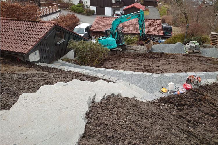 Grønn gravemaskin arbeider på en skråning med steintrapper og nyanlagt hage i bakgrunnen.