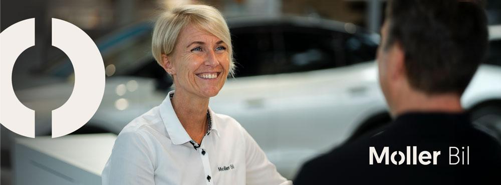 Kvinne med kort hår smiler mens hun snakker med en kunde i en bilforhandler. Møller Bil-logo synlig.