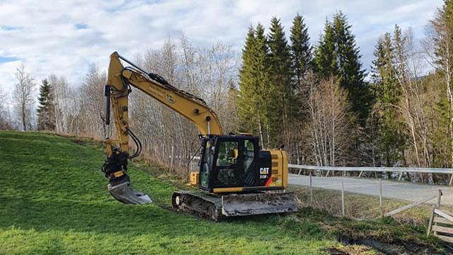 Gravemaskin i arbeid på en grønn bakke ved veien, omgitt av trær og en klar himmel.