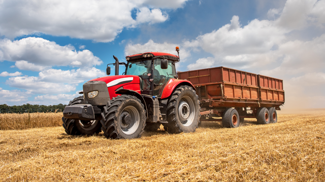 Rød traktor som trekker en vogn gjennom et kornfelt under en blå himmel med skyer.