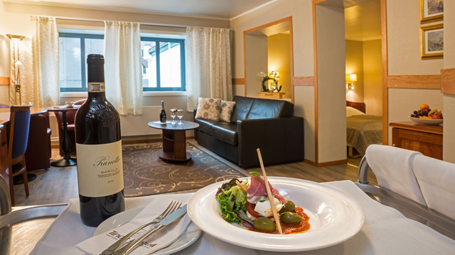 Et bord med en tallerken med mat, en flaske vin og en stue med sofa og vindu i bakgrunnen.