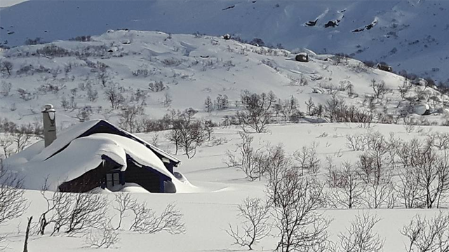 Et koselig hus dekket av snø i et vinterlandskap med trær og fjell i bakgrunnen.