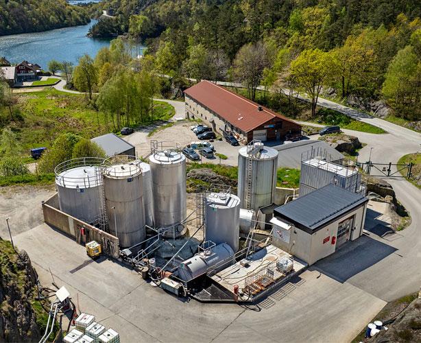Oversiktsbilde av en industriell anlegg med tanker og bygninger ved en innsjø omkranset av grønn natur.