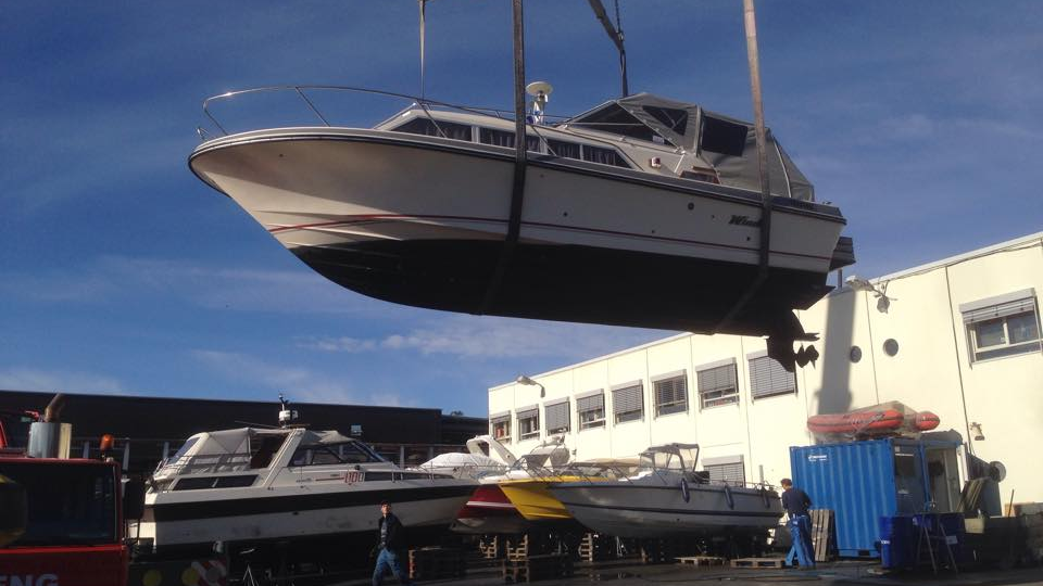 Båt heises opp av en kran ved en marina, med flere båter og bygninger i bakgrunnen.
