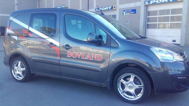 Grå varebil med Dovland-logo og dekorative striper parkert foran en bygning.