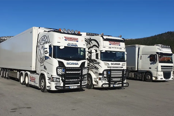 Tre lastebiler fra Scania parkert på en vei med fjell i bakgrunnen. Lastebilene har hvit farge og dekorative detaljer.