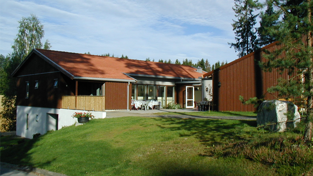 Moderne enebolig med rød takstein, terrasse og store vinduer, omringet av trær og grønt gress.