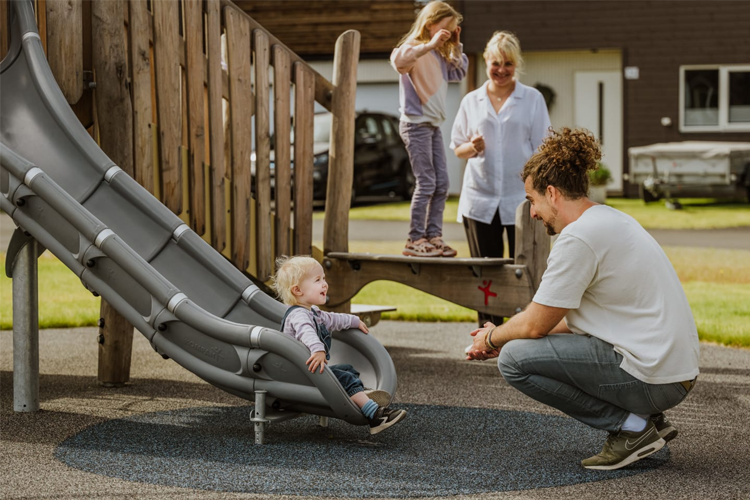 Barn leker på lekeplass med sklie, en voksen sitter på bakken og smiler til et lite barn som kommer ned fra sklien.