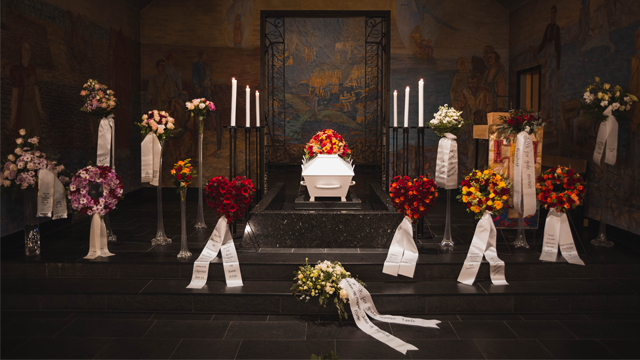 En kiste omgitt av blomsterkranser og lys i et kapell, med malerier på veggene.