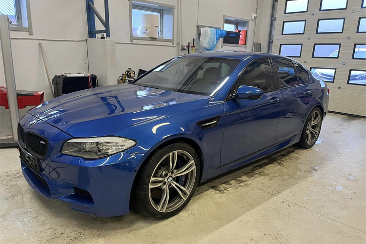 Blå BMW M5 parkert i en garasje med lys og verktøy i bakgrunnen.