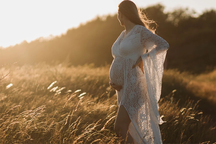 Gravid kvinne i hvit kjole står i solnedgang på en eng med høyt gress.