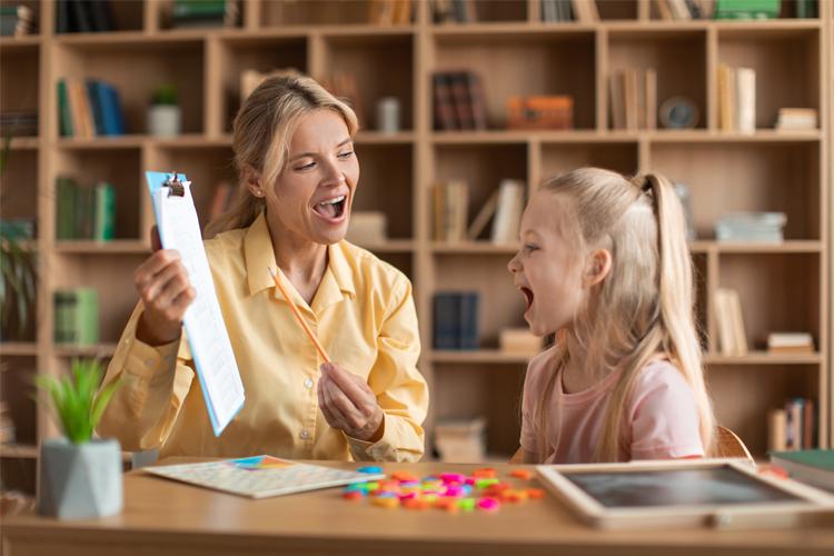 En voksen kvinne og en jente sitter ved et bord, smilende og syngende mens de holder på med kreative aktiviteter.