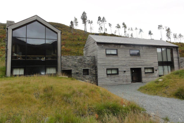 Moderne hus med store vinduer og steinmur, omringet av grønt landskap og trær i bakgrunnen.