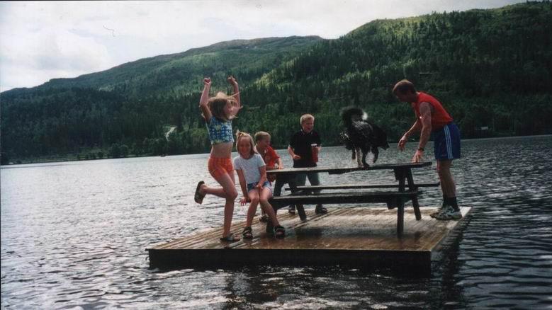 Barn som leker på en flytende plattform i en innsjø, med fjell i bakgrunnen. Solfylt dag med glede og aktivitet.