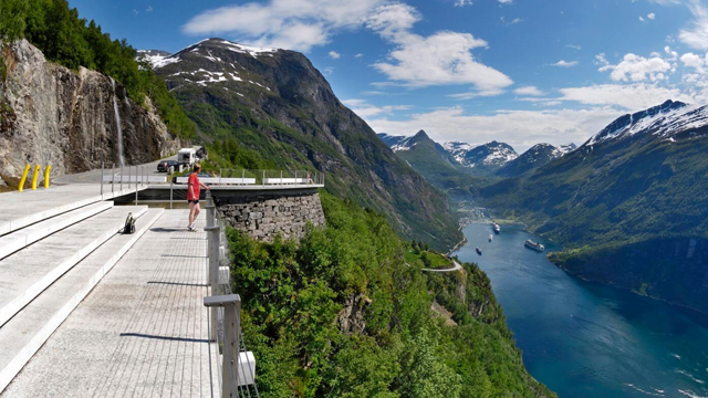 Utsikt over fjorden fra en utsiktsplattform med fjell og grønn vegetasjon i bakgrunnen.