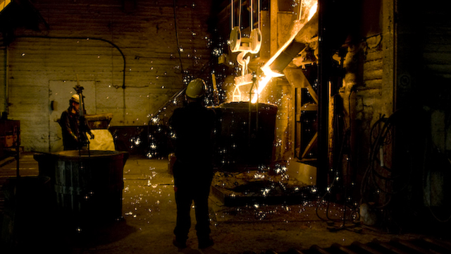 En arbeider i hjelm står foran en smelteovn, mens gnister og bobler flyr i luften i en smeltefabrikk.