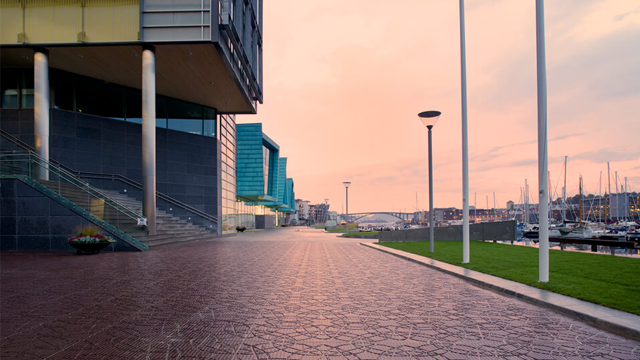 Moderne havnepromenade med bygninger, gatebelysning og båter i bakgrunnen under en rosa himmel.