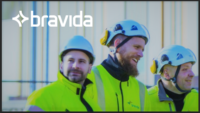 Tre arbeidere i gule sikkerhetsjakker og hjelmer smiler mens de står sammen på en byggeplass.