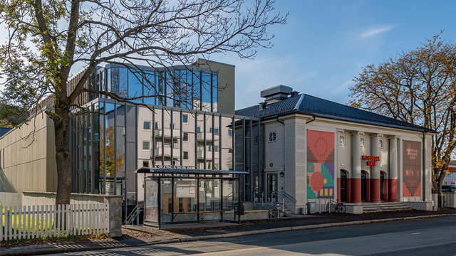 Moderne bygning med glassfasade ved siden av en klassisk bygning med søyler og kunstverk på veggen.
