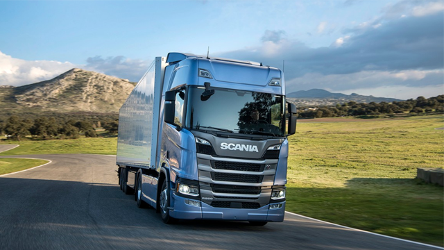 En blå Scania lastebil kjører på en vei med grønne åser i bakgrunnen.