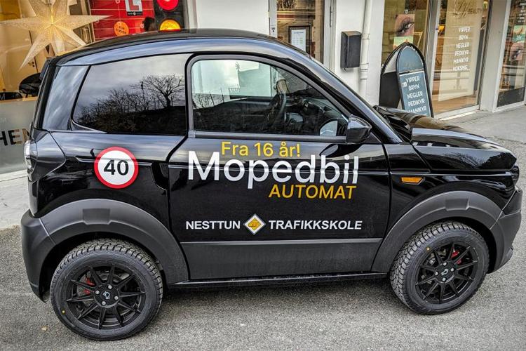 Svart mopedbil med tekst om automatkjøring og aldersgrense, parkert ved trafikkskole.