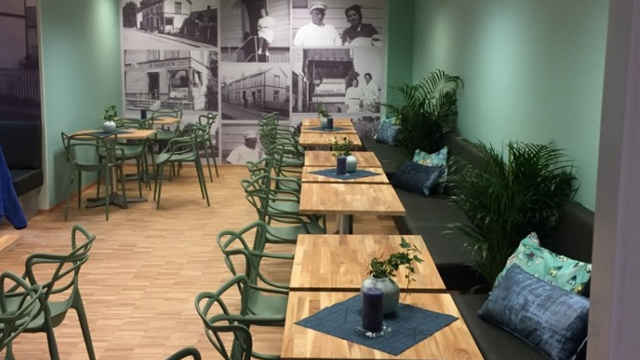 Moderne kaféinnredning med grønne vegger, trebord og planter, samt bilder på veggen som viser historiske scener.