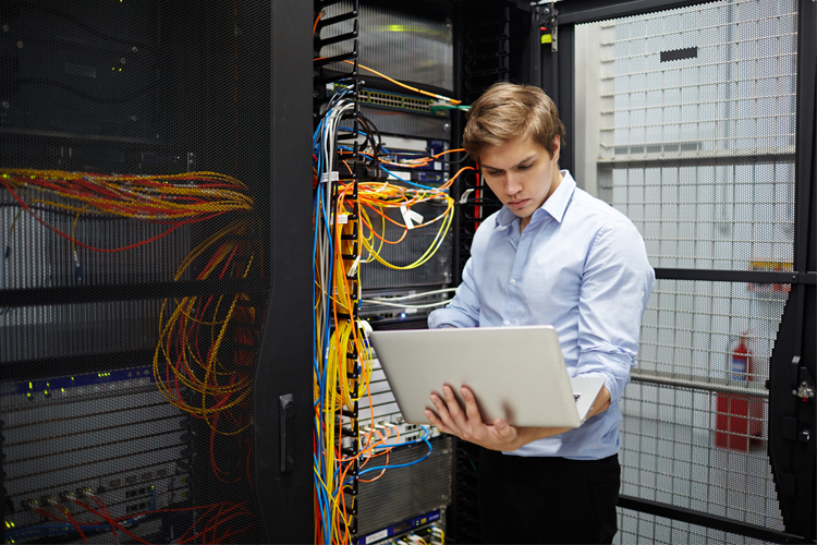 En ung mann jobber med en bærbar datamaskin i et serverrom med mange kabler.