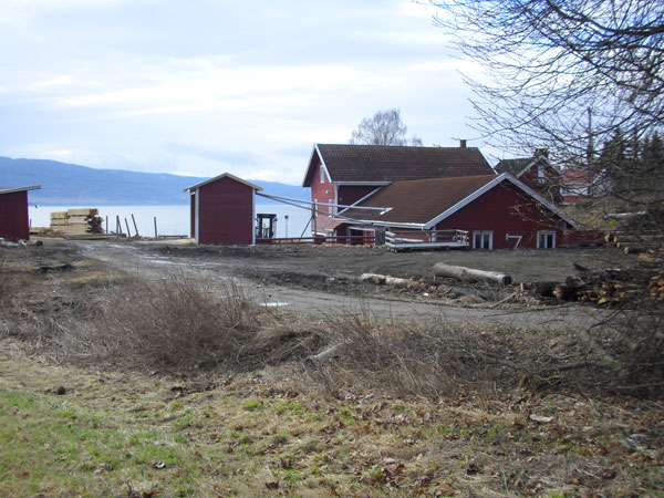 Rødt hus med uthus ved vannet, omgitt av trær og en grusvei i forgrunnen.